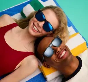 Two people lying down seen from above: a man with light brown skin and a woman with light skin and blonde hair. Both are wearing sunglasses with unisex injected plastic frames. The man's lenses are polarized and black, the woman's are polarized and blue.