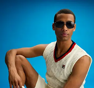 A man with light brown skin is wearing sunglasses with dark lenses and a metal frame. He is seen from the front, looking at the camera, and the background is a solid light blue.