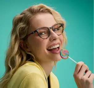 Una donna con pelle chiara, capelli biondi e mossi, vista di profilo (parte superiore del corpo), sorride mentre lecca un lecca-lecca. Indossa occhiali da vista neri con logo FILA sulle aste.