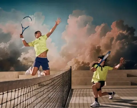 Two athletes are playing tennis on an outdoor court with a dramatic cloudy sky in the background. Both wear matching sports outfits, consisting of yellow shirts, black shorts, and tennis shoes. One player is airborne, about to hit the ball with a racket, while the other is in a defensive stance, ready to respond. The image conveys action, competition, and dynamism.