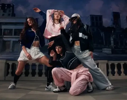 Four young people pose in urban sportswear on a terrace with a city backdrop. They wear casual items such as wide pants, shorts, sporty jackets, crop tops, and sneakers. Their poses are dynamic and choreographed, suggesting a group dance or street dance context. The atmosphere is energetic and modern, highlighting style and coordination among the group members.